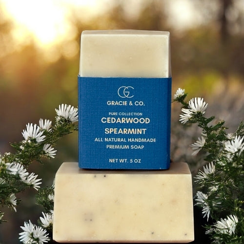 Bar of soap with a blue label on a wooden block, surrounded by greenery and flowers.